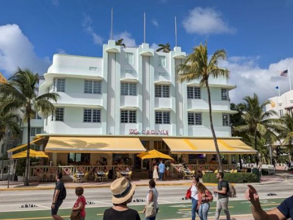 A street scene with people walking and a large Art Deco-style building, palm trees, and a restaurant with a yellow canopy are visible.
