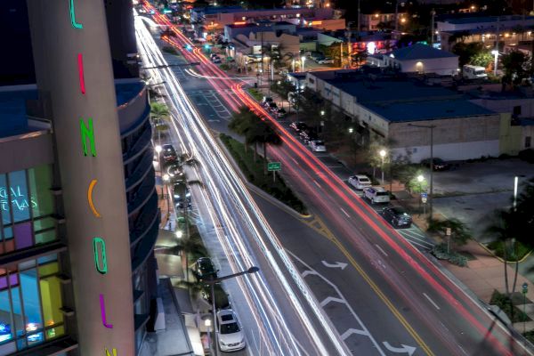 A nighttime city scene with light trails from cars along a busy road. The building on the left has colorful vertical signage spelling "LINCOLN."