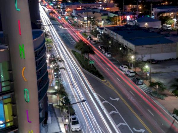A nighttime city scene with light trails from cars along a busy road. The building on the left has colorful vertical signage spelling "LINCOLN."