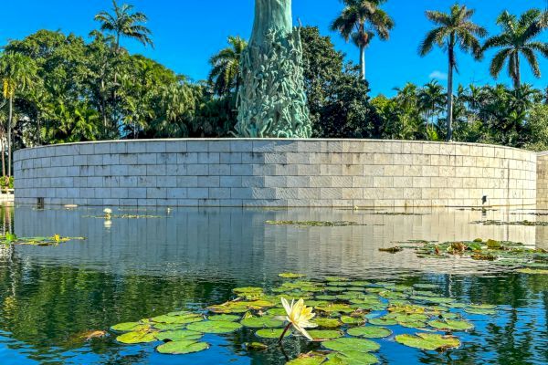 The image shows a statue surrounded by a circular stone wall and a pond with lily pads, with tall palm trees and lush greenery in the background.