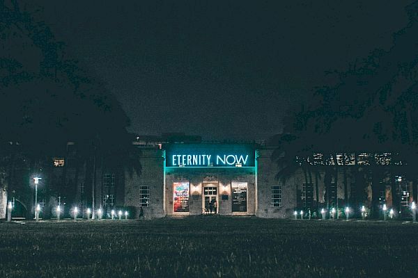 A building with a neon sign that reads "ETERNITY NOW" is illuminated at night, surrounded by trees and outdoor lighting.