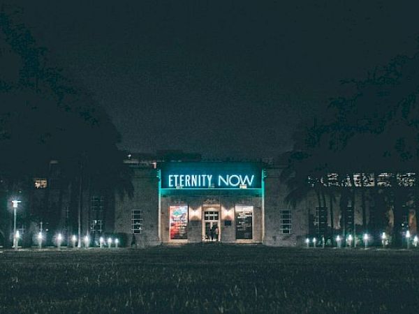 A building with a neon sign that reads "ETERNITY NOW" is illuminated at night, surrounded by trees and outdoor lighting.