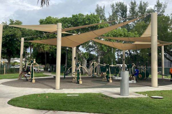 An outdoor fitness area with exercise equipment is sheltered by beige canopies in a park setting with surrounding greenery.