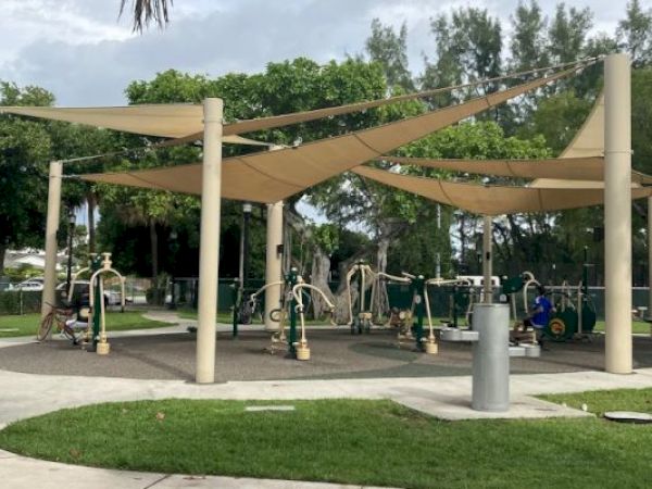 An outdoor fitness area with exercise equipment is sheltered by beige canopies in a park setting with surrounding greenery.