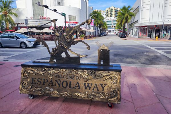 The image shows a decorative sign that reads "Welcome to Española Way" with a statue of a historical figure, set on a pink tiled street.