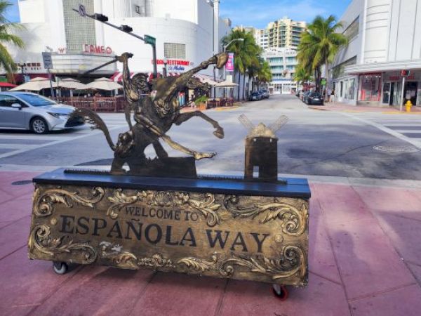The image shows a decorative sign that reads "Welcome to Española Way" with a statue of a historical figure, set on a pink tiled street.