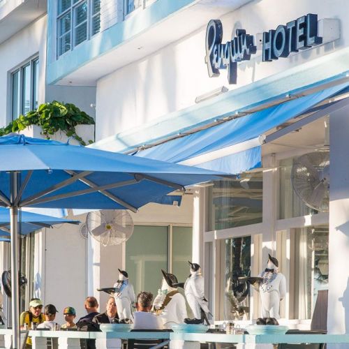The image shows an outdoor cafe with blue umbrellas and the sign "Penguin Hotel" on the building. People are seated at tables enjoying a meal.