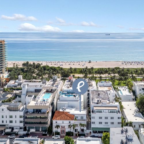 A cityscape with buildings, palm trees, a pool, and a beach next to the ocean under a blue sky.