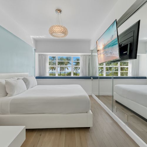 The image shows a modern bedroom with a king-sized bed, a mirrored wall, a wall-mounted TV, and a window with a view of palm trees outside.