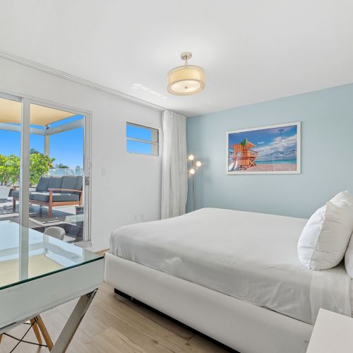 A modern bedroom with a large bed, desk, lamp, and wall art, leading to a balcony with seating and a view of blue skies.