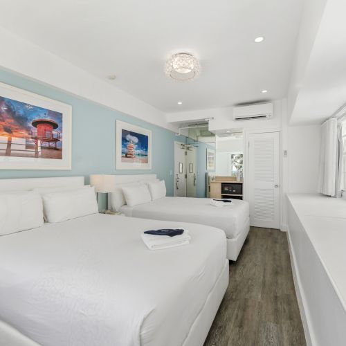 A bright hotel room features two beds, blue walls, beach-themed artwork, and a mounted TV, creating a relaxing atmosphere.