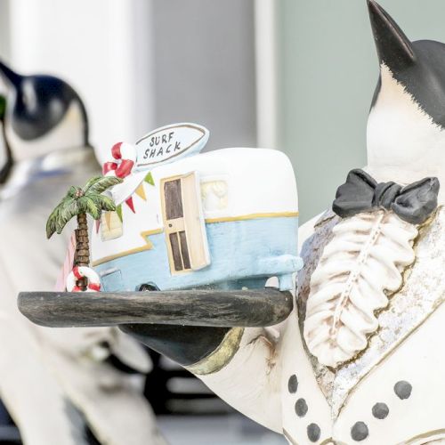 A penguin statue in a tuxedo holds a miniature "surf shack" with a palm tree. Other similarly dressed penguin statues are in the background.