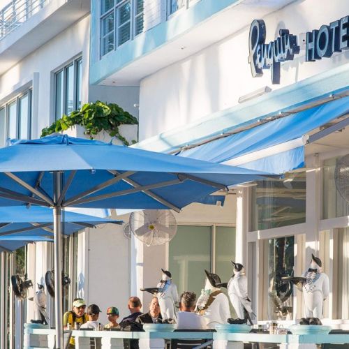 This image shows the exterior of a hotel with a blue awning and outdoor seating area occupied by people beneath blue umbrellas, labeled "Penguin Hotel."