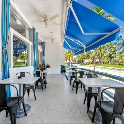 Outdoor cafe with tables, chairs, and blue awnings, overlooking a sunny, palm tree-lined street.