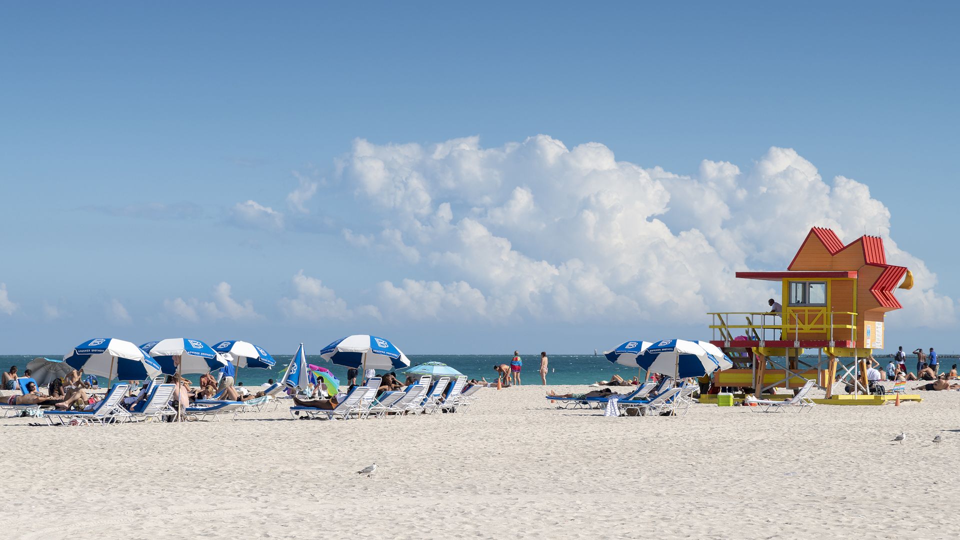 People relax under blue and white umbrellas on a sandy beach with a colorful lifeguard tower and ocean in the background under a clear sky.