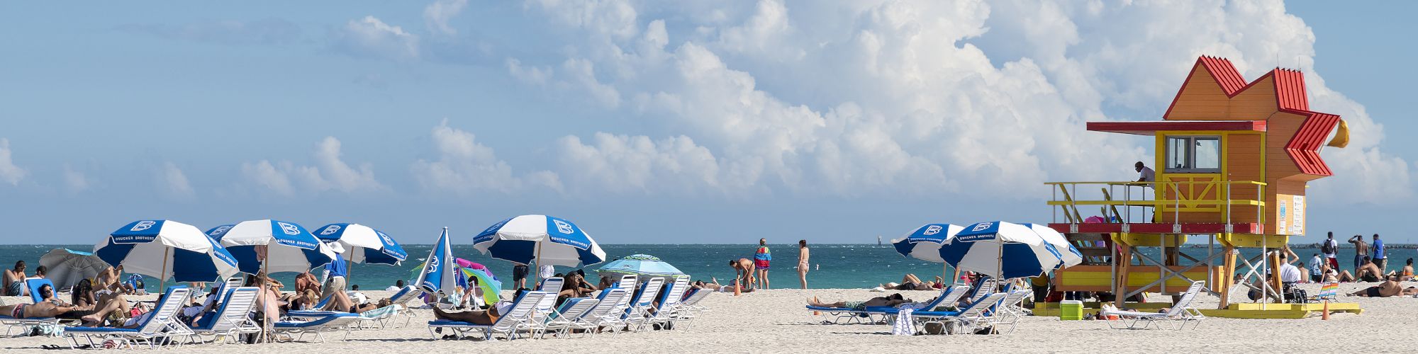 People relax under blue and white umbrellas on a sandy beach with a colorful lifeguard tower and ocean in the background under a clear sky.