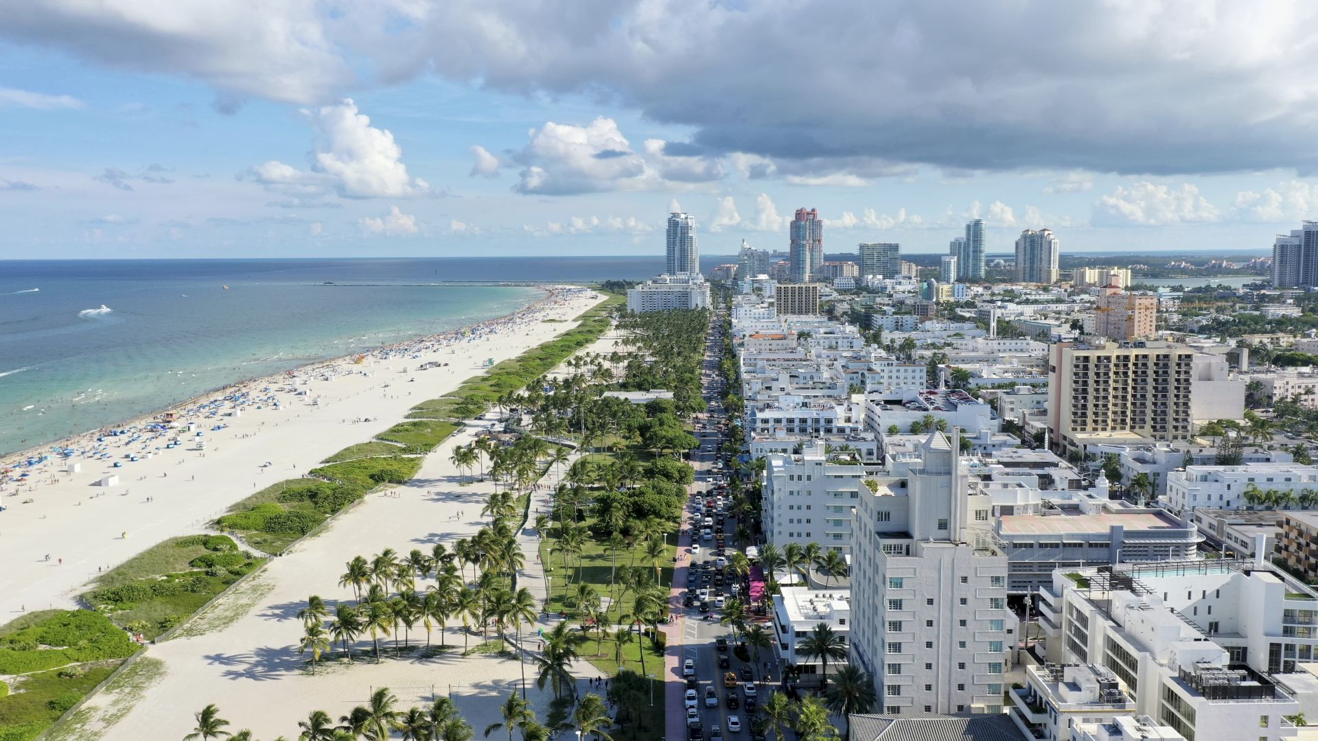 An aerial view of a beachfront city with high-rise buildings, a crowded beach, lush green palm trees, and a clear sky with scattered clouds ends the sentence.