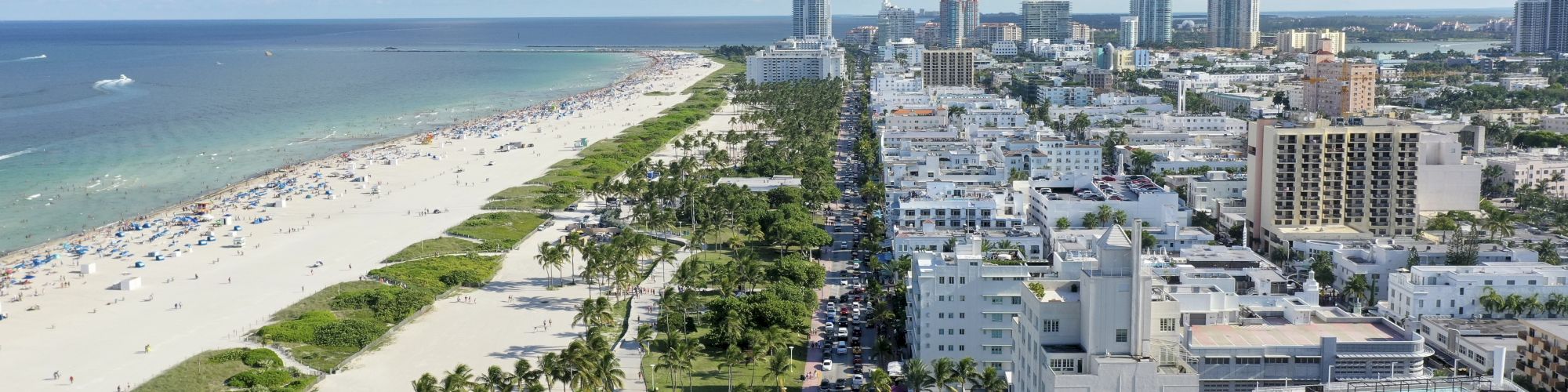 An aerial view of a beachfront city with high-rise buildings, a crowded beach, lush green palm trees, and a clear sky with scattered clouds ends the sentence.