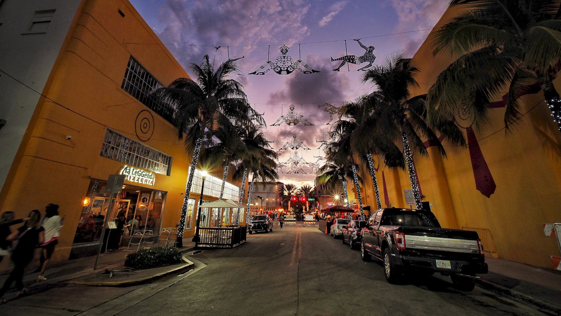 A twilight street scene with lit palm trees, yellow buildings, parked cars, and string lights overhead, creating a festive atmosphere.