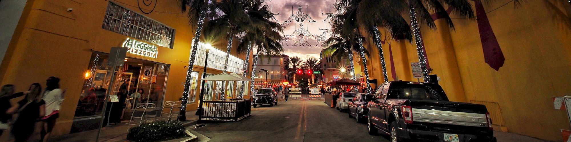 A twilight street scene with lit palm trees, yellow buildings, parked cars, and string lights overhead, creating a festive atmosphere.