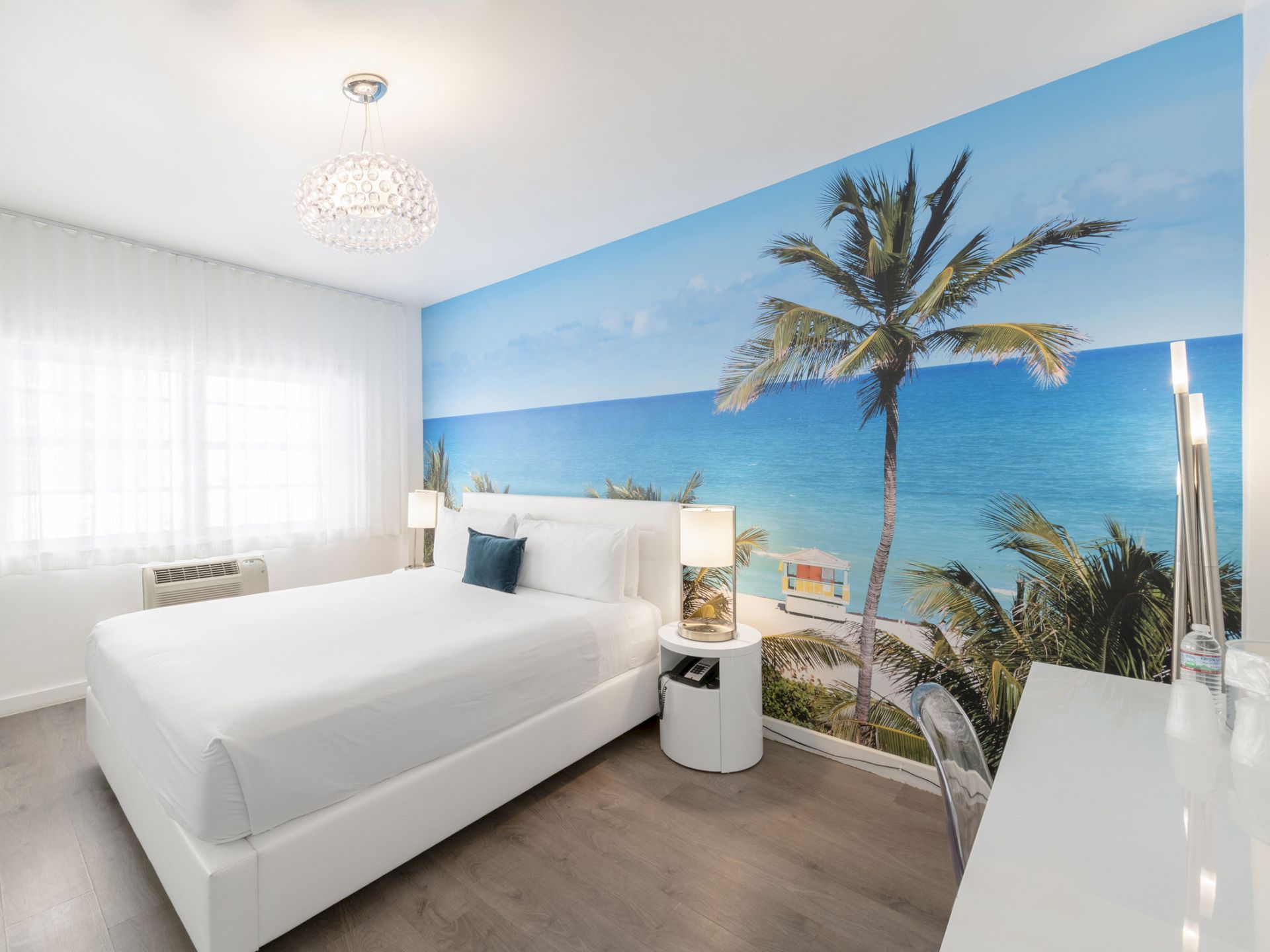A white hotel room with a large beach mural on one wall, a king-size bed, and a chandelier.