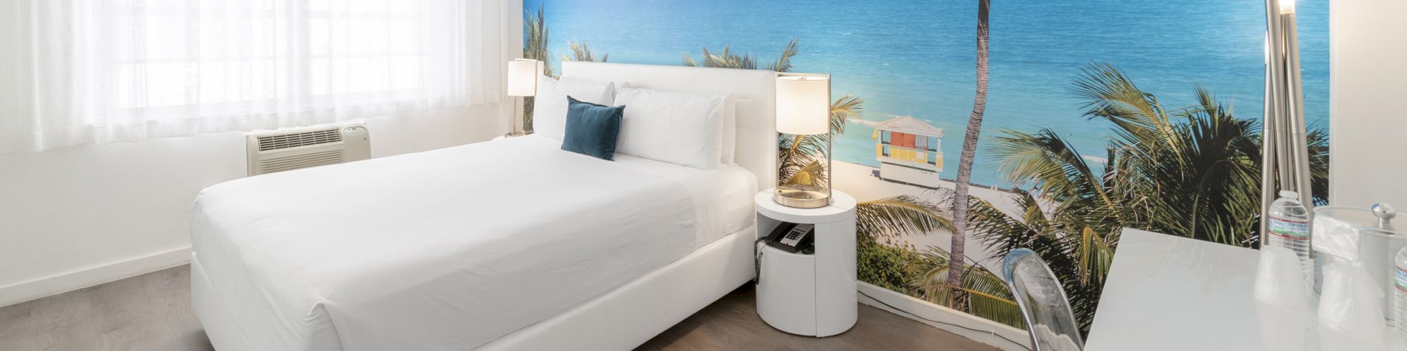A modern bedroom with a beach mural wall, featuring a white bed, bedside tables, and a chandelier. The room has large windows and wooden flooring.