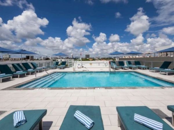 A rooftop swimming pool with lounge chairs and umbrellas under a blue sky with fluffy clouds. Towels are neatly placed on the lounge chairs.