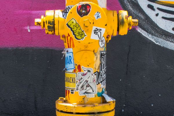 A yellow fire hydrant covered in stickers is situated in front of a graffiti wall with a mix of black, white, and pink colors ending the sentence.