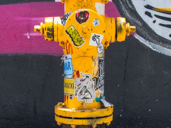 A yellow fire hydrant covered in stickers is situated in front of a graffiti wall with a mix of black, white, and pink colors ending the sentence.