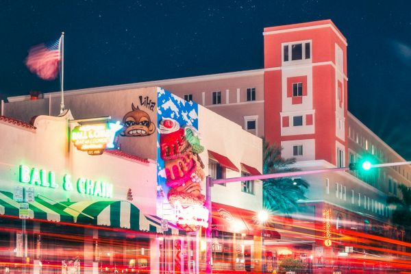 A vibrant cityscape at night with a neon-lit building, light trails from cars, a starry sky, and a prominent red structure in the background.