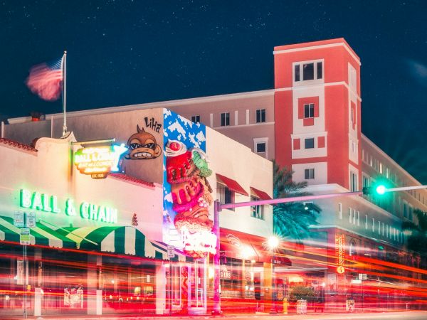 A vibrant cityscape at night with a neon-lit building, light trails from cars, a starry sky, and a prominent red structure in the background.