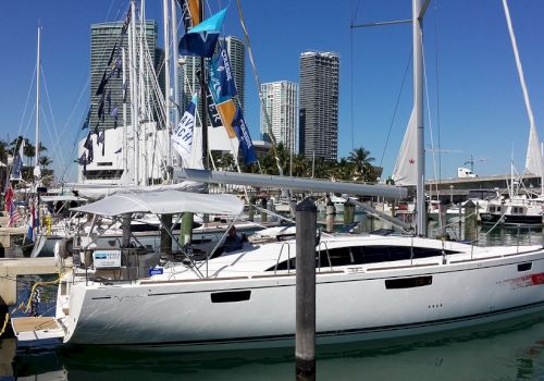 A modern sailboat docked at a marina with city buildings in the background and clear blue skies above, surrounded by other boats.