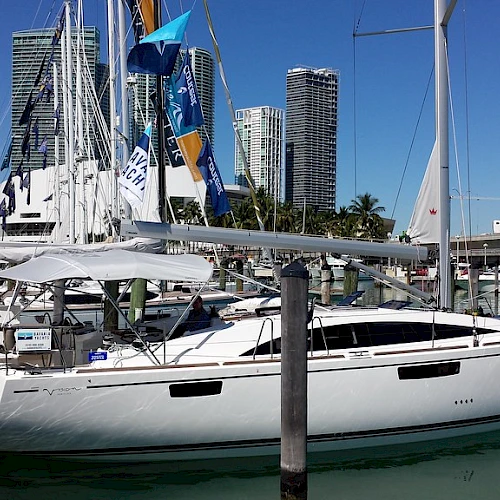 A modern sailboat docked at a marina with city buildings in the background and clear blue skies above, surrounded by other boats.