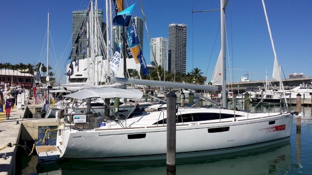 A modern sailboat docked at a marina with city buildings in the background and clear blue skies above, surrounded by other boats.