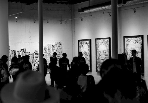 A group of people in a dimly lit gallery observes large framed artworks on the walls, creating a vibrant and thought-provoking atmosphere.