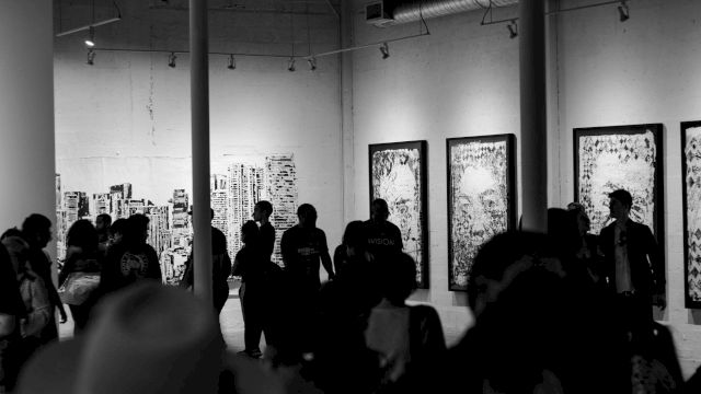 A group of people in a dimly lit gallery observes large framed artworks on the walls, creating a vibrant and thought-provoking atmosphere.