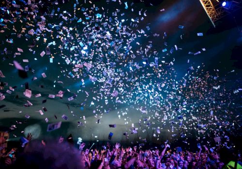A crowd enjoys a concert under blue lights with confetti raining down, creating a lively and festive atmosphere.