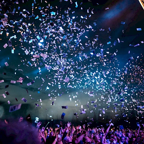 A crowd enjoys a concert under blue lights with confetti raining down, creating a lively and festive atmosphere.