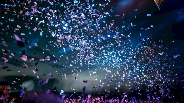 A crowd enjoys a concert under blue lights with confetti raining down, creating a lively and festive atmosphere.