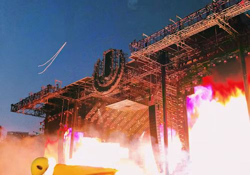 A vibrant music festival scene with a large stage, bright lights, and a crowd waving a flag under a clear sky at sunset.
