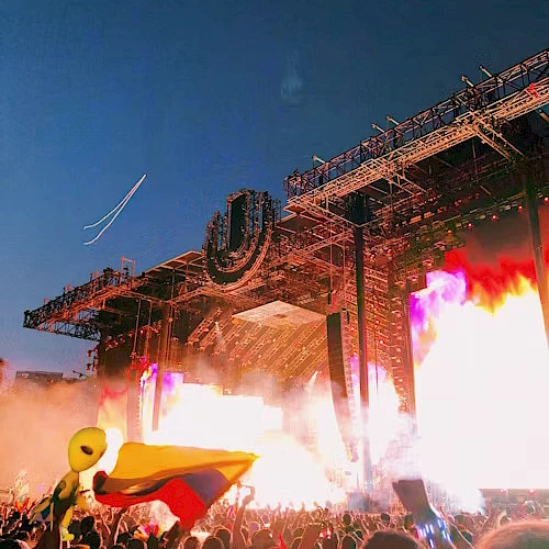 A vibrant music festival scene with a large stage, bright lights, and a crowd waving a flag under a clear sky at sunset.