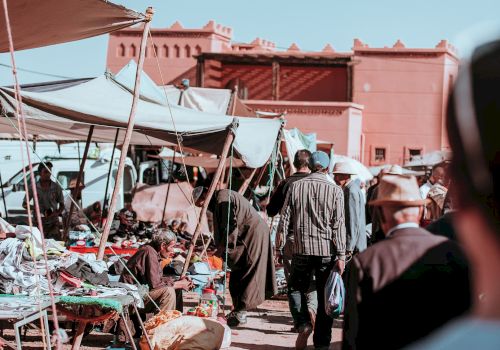 A bustling outdoor market scene with people walking, vendor stalls, and a building in the background against a clear sky.