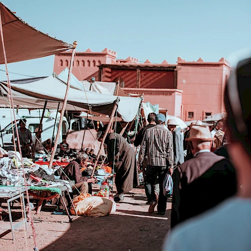 A bustling outdoor market scene with people walking, vendor stalls, and a building in the background against a clear sky.