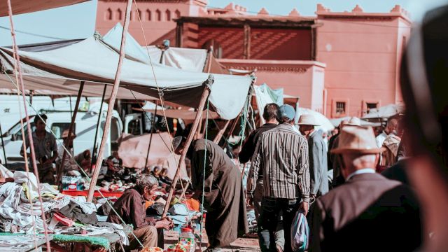 A bustling outdoor market scene with people walking, vendor stalls, and a building in the background against a clear sky.