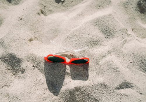 A pair of red sunglasses and two straws are lying on sandy beach with sunlight casting shadows, creating a relaxed, summery vibe.