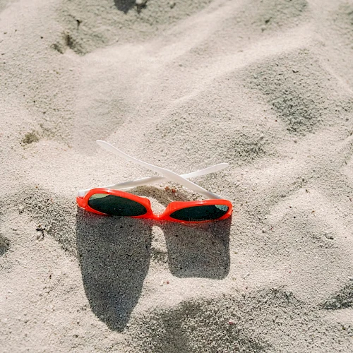 A pair of red sunglasses and two straws are lying on sandy beach with sunlight casting shadows, creating a relaxed, summery vibe.
