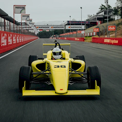 A yellow race car drives on a track with red barriers and advertising signs, under a cloudy sky. The focus is on the vehicle's front view.