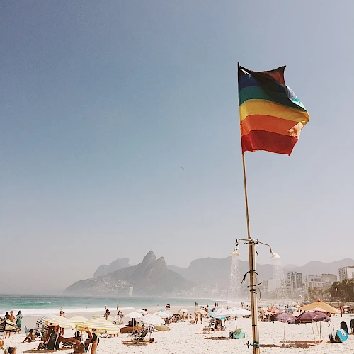 A beach scene with people relaxing on the sand and a multicolored flag on a pole, with mountains visible in the background.