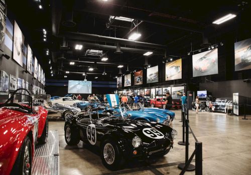 The image shows a car museum exhibit with classic cars, including a black one labeled "99," surrounded by visitors and automotive displays.