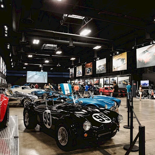 The image shows a car museum exhibit with classic cars, including a black one labeled "99," surrounded by visitors and automotive displays.